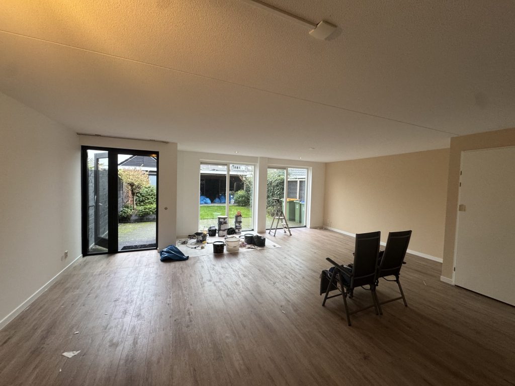 Ruime woonkamer met lichte wanden, openslaande deuren naar de tuin en verfspullen op de vloer